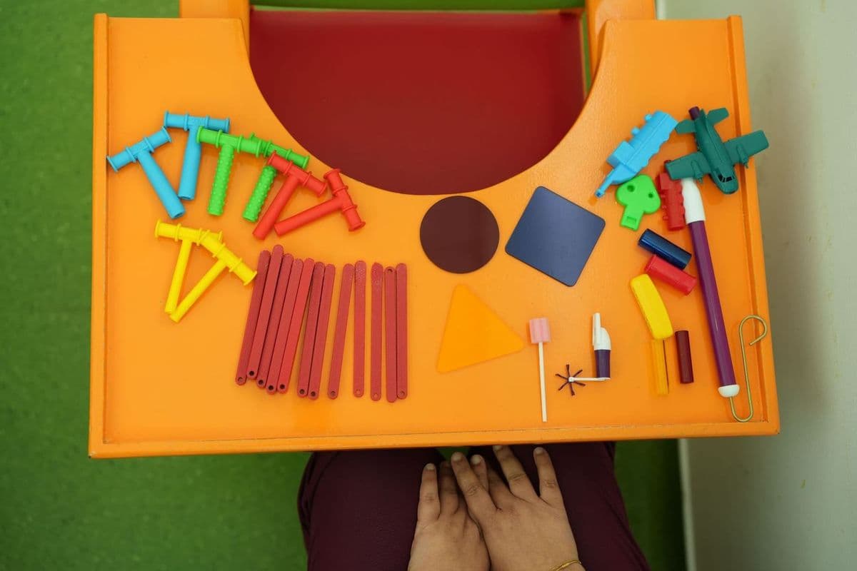 An overhead view of a child's orange therapy tray filled with colorful developmental toys, including textured Chewy Tubes, wooden sticks, geometric shapes, and sensory tools.