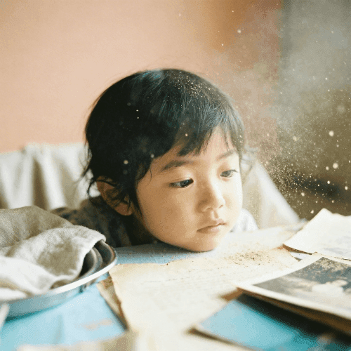 Close-up of a young child with dark hair resting their chin near a table, watching sparkling gold dust float through the air in warm, soft light.