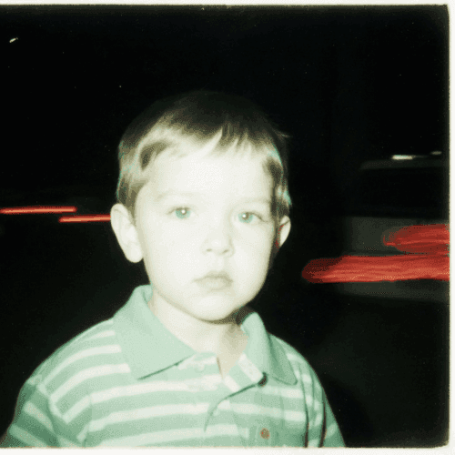 A young child in a striped shirt looks forward calmly against a dark background with blurred red lights shining behind.