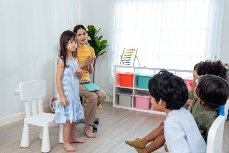 A young girl speaking in front of a classroom circle while a teacher and other children listen attentively.